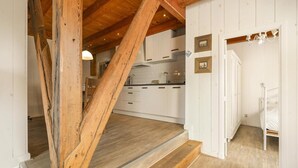 Un intérieur rustique avec des poutres apparentes, une cuisine dotée de meubles blancs et un escalier menant à une chambre à coucher où se trouve un lit blanc.