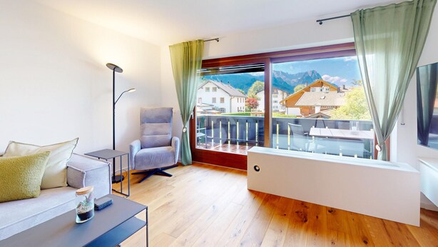 Un salon moderne comprenant un canapé, un fauteuil, une table basse et une grande fenêtre donnant sur des maisons et des montagnes.
