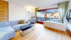 Un salon moderne avec un canapé, une table basse et un fauteuil. Une grande fenêtre offre une vue sur les bâtiments et les montagnes.