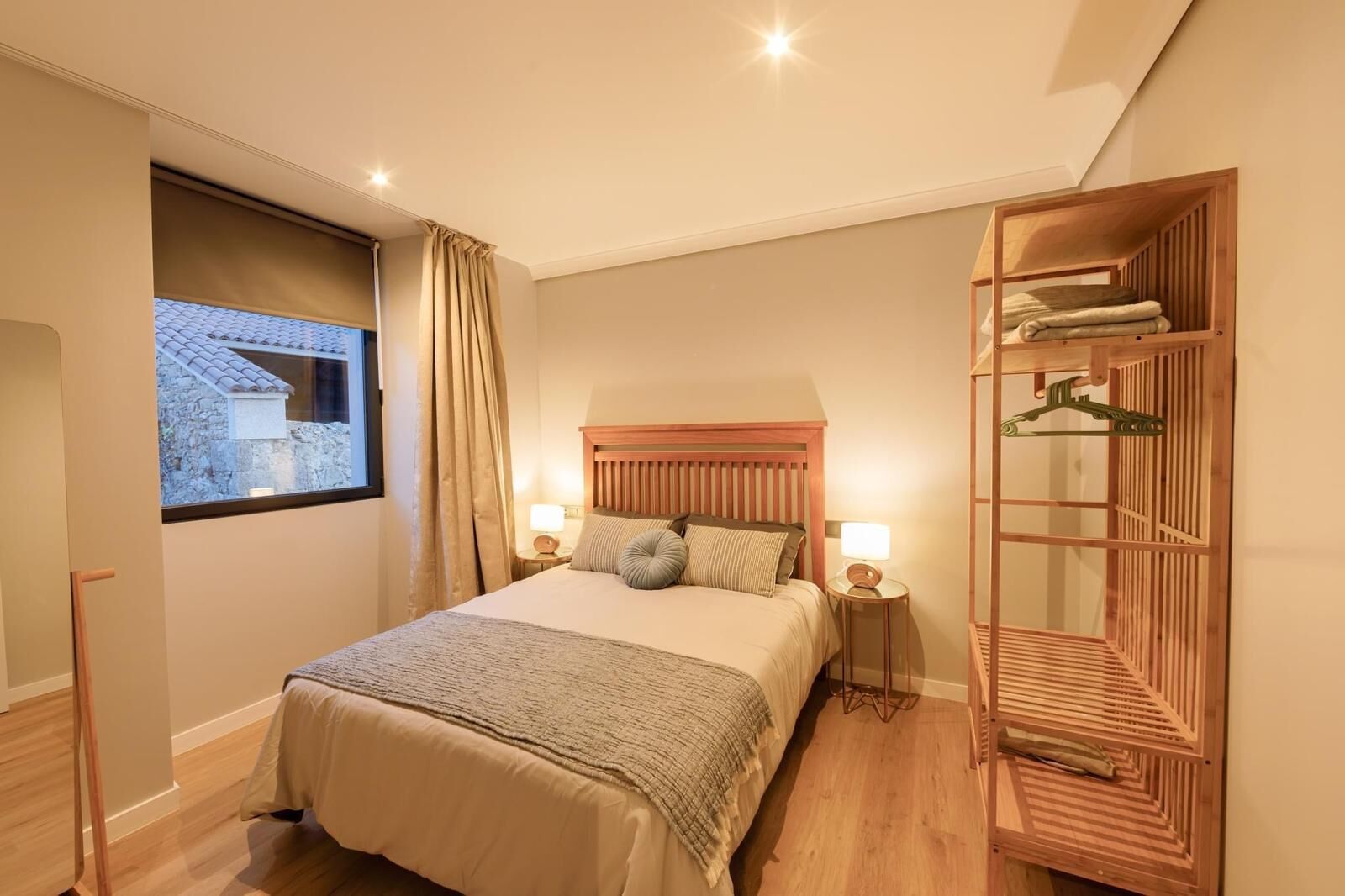 A bedroom with a bed, bedside lamps, a window with curtains, a wooden wardrobe, and a view of a building outside.