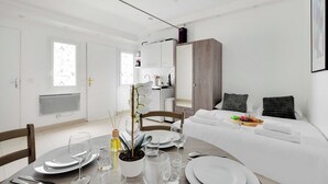 Une pièce moderne et bien éclairée, avec une table à manger dressée pour quatre personnes, un canapé agrémenté de coussins et un lit sur lequel se trouve un plateau de fruits.