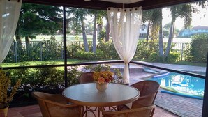 A covered outdoor seating area with wicker chairs and a table, a small flower vase, and a view of a swimming pool and greenery. - PGA National Luxury Estate Home, Golf and Lake, Discount @ 3+ weeks (Palm Beach Gardens)