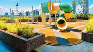 A playground with a slide and climbing structures, surrounded by planters and a paved area.