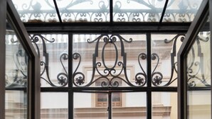 Uitzicht door een ijzeren hekwerk met een balkon, leidend naar een gebouw met een witte gevel en houten luiken.