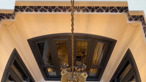 Een kroonluchter met decoratieve voet, een rond wandornament en een raam met uitzicht op de lucht.