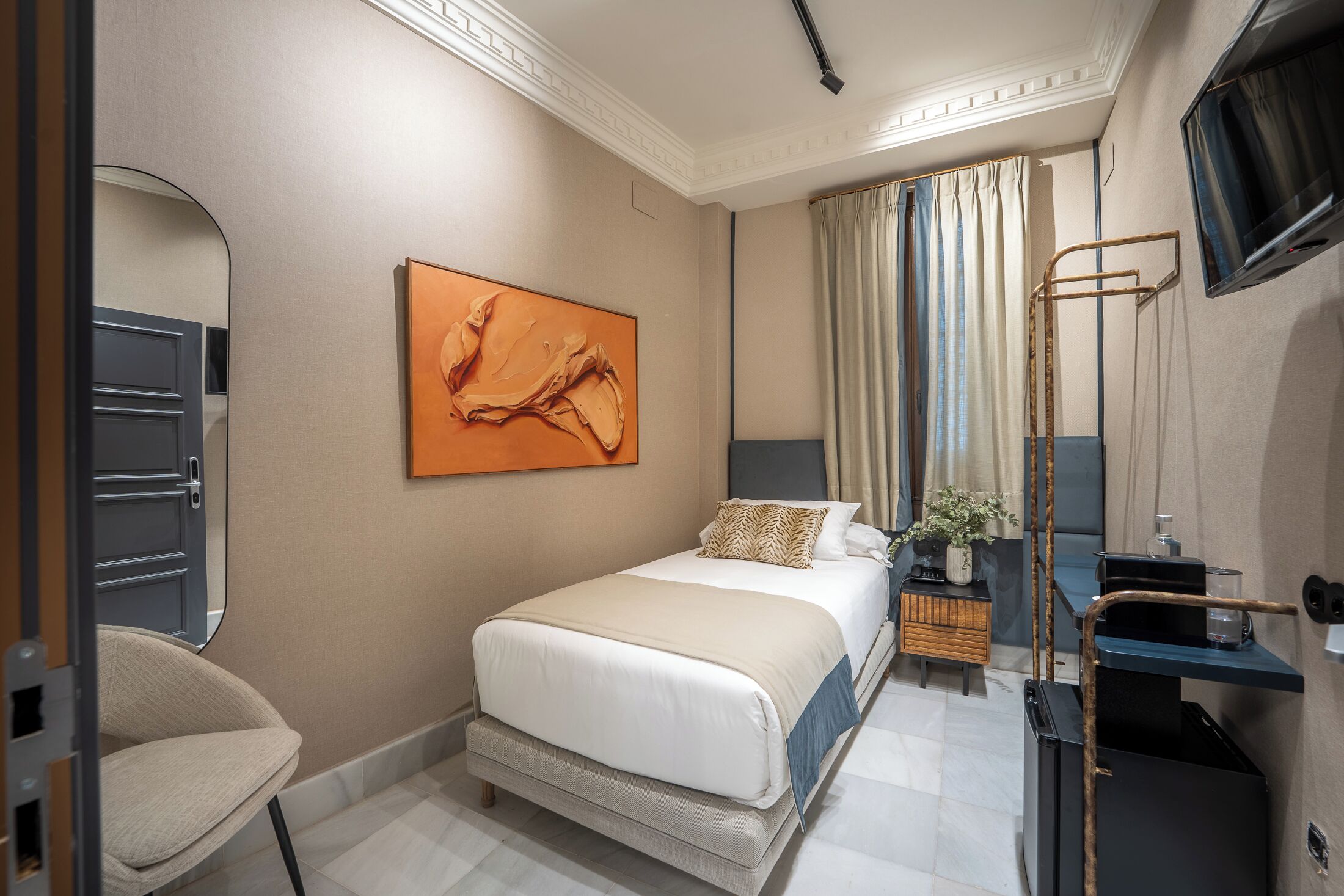 A hotel room with a bed, a chair, a desk, a TV, and a painting on the wall.
