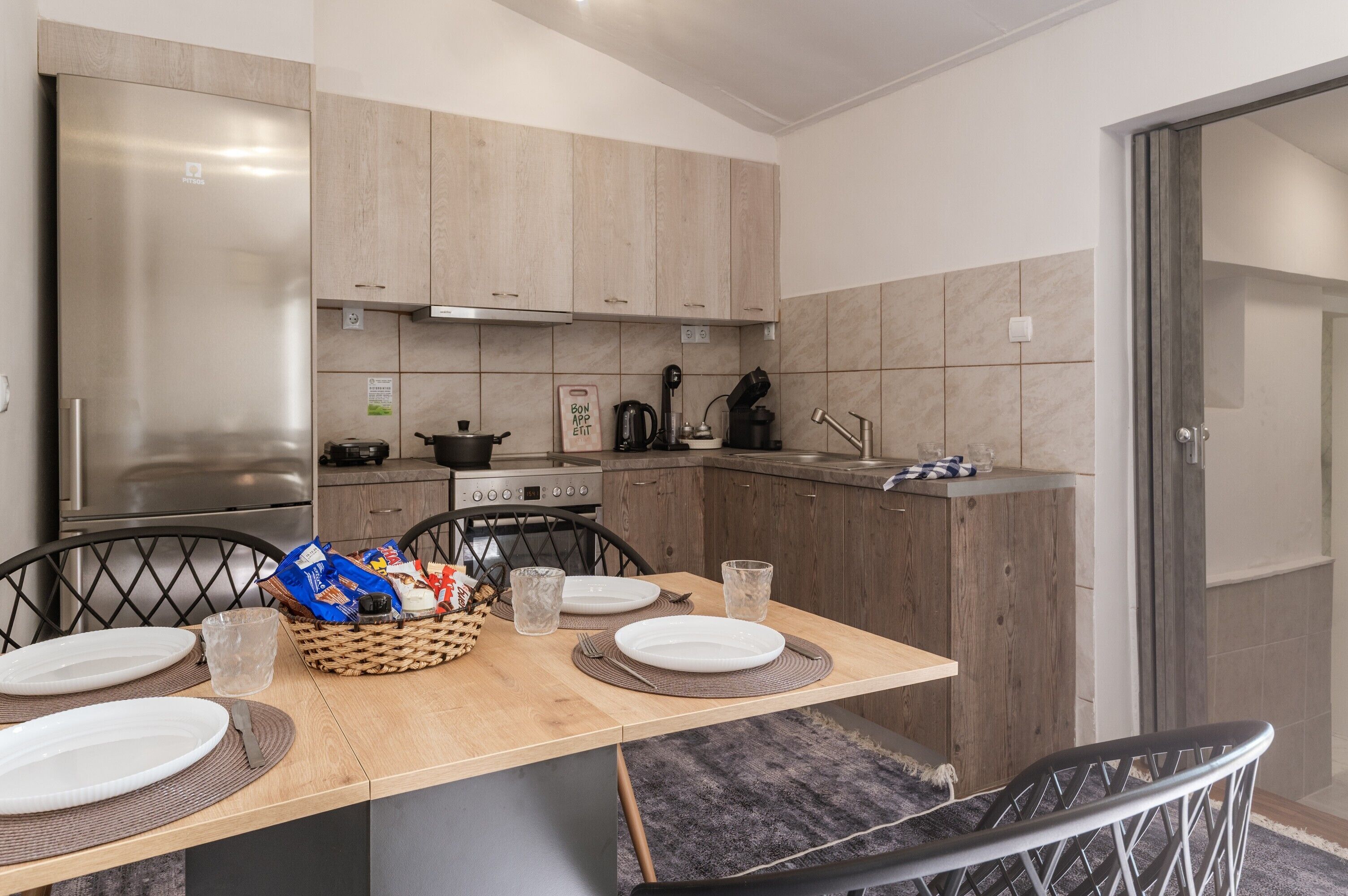 A modern kitchen with wooden cabinets, a stainless steel refrigerator, a dining table set with plates, glasses, and a basket of snacks.