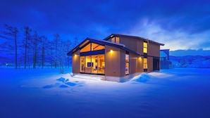 A modern house with large windows and a well-lit interior, surrounded by snow. - Sakuraya (Hakuba)