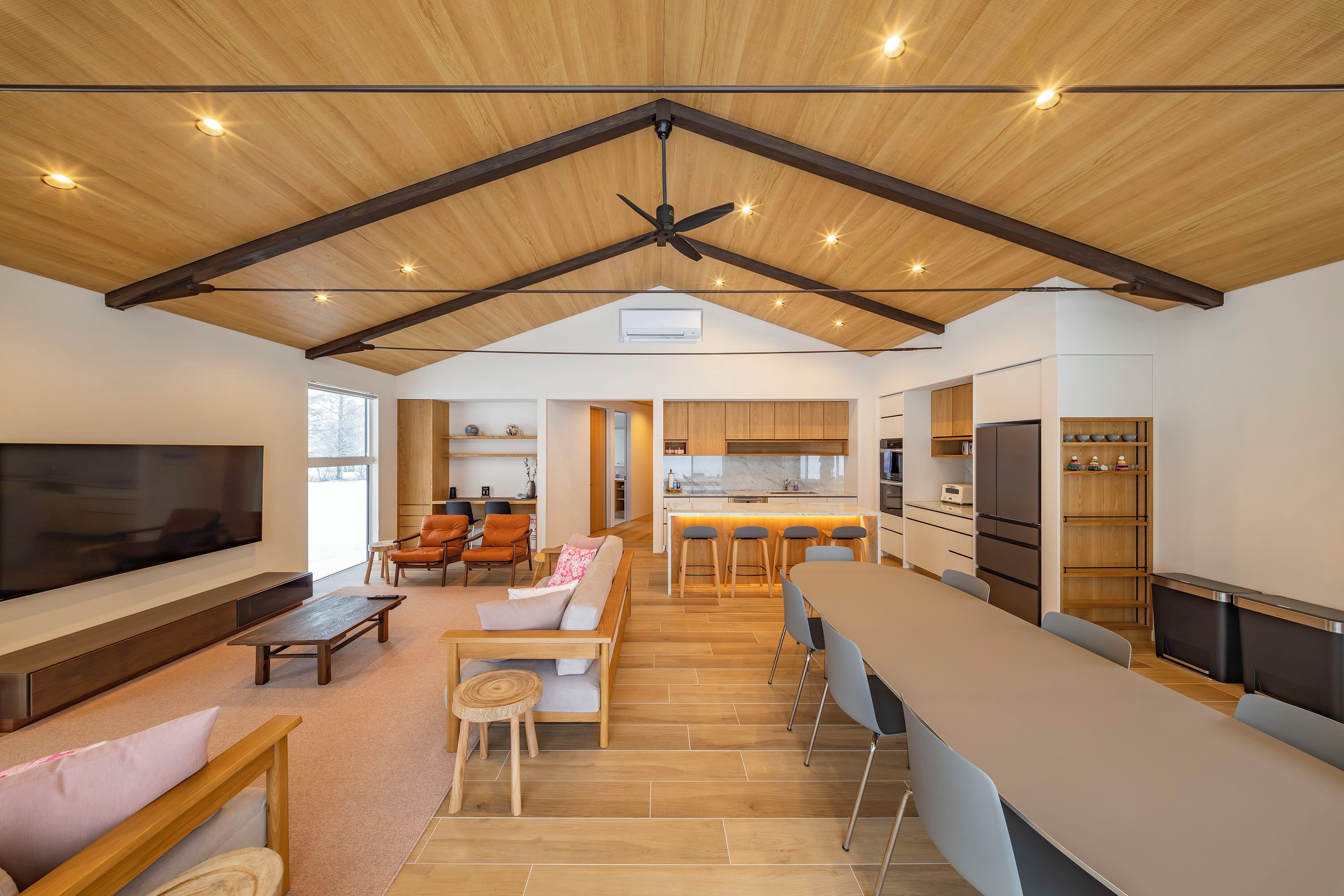 A spacious living area with a large TV, wooden furniture, and a kitchen in the background.