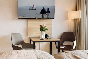 A hotel room with two chairs, a small table with a vase of flowers, a TV displaying a scenic view, and a lamp.