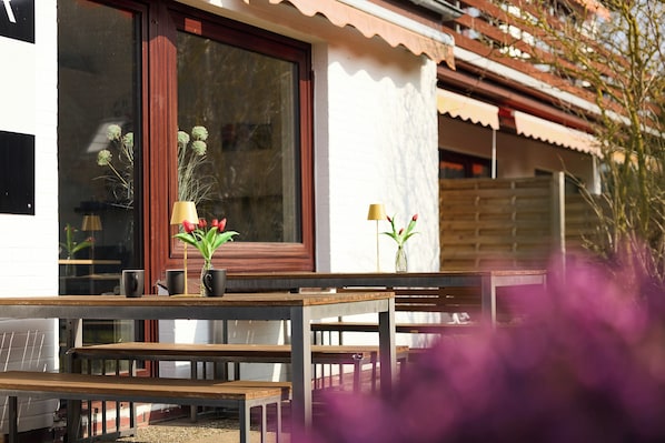 A wooden outdoor seating area with tables and benches, a window with a view, and a small vase with flowers.