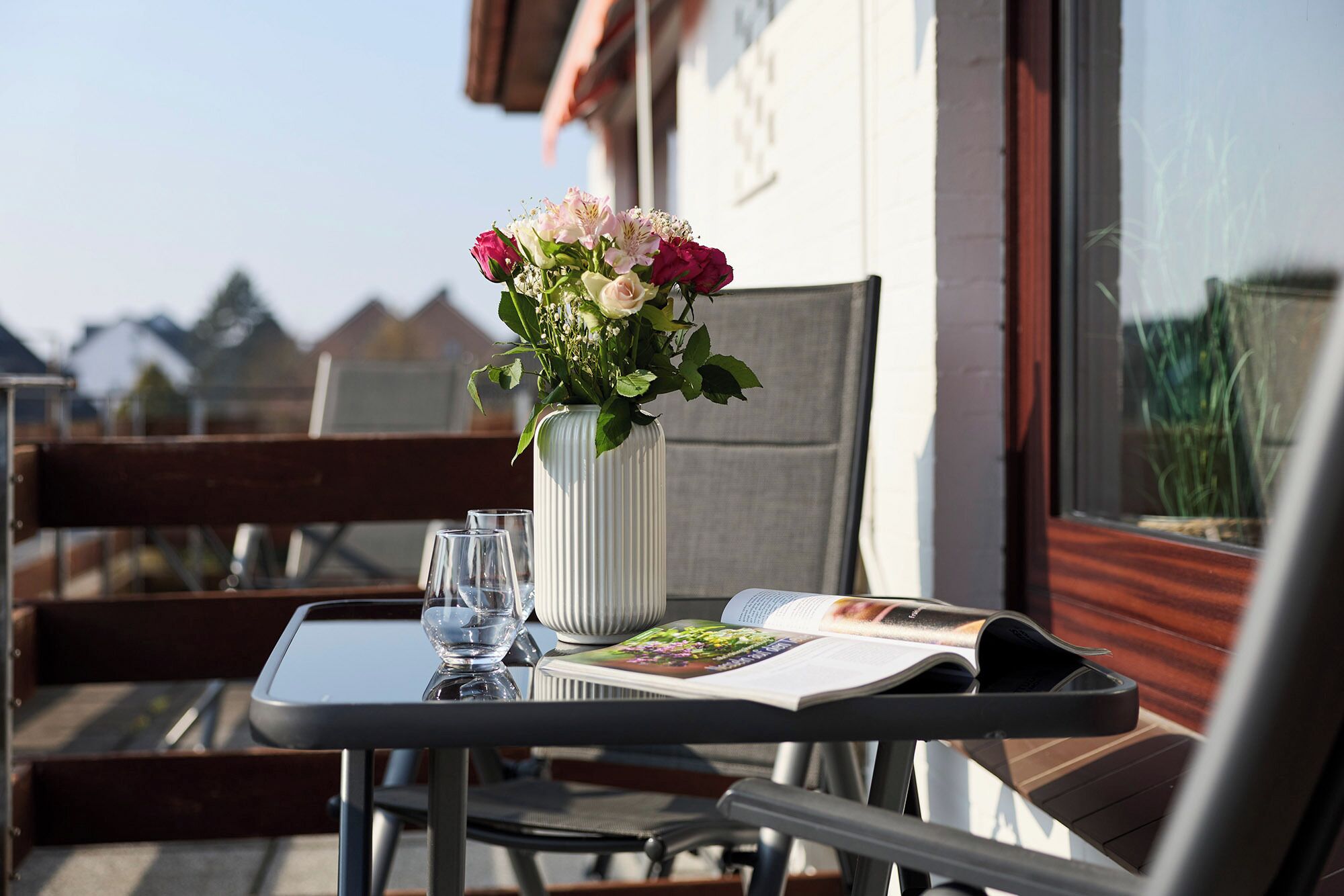 Et bord med en vase blomster, to glas og et magasin på en balkon.
