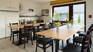 A dining area with wooden tables and black chairs, a kitchen in the background, and a view through large windows. - Quartier 7 apartment in a quiet location and access to the sea time (Büsum)