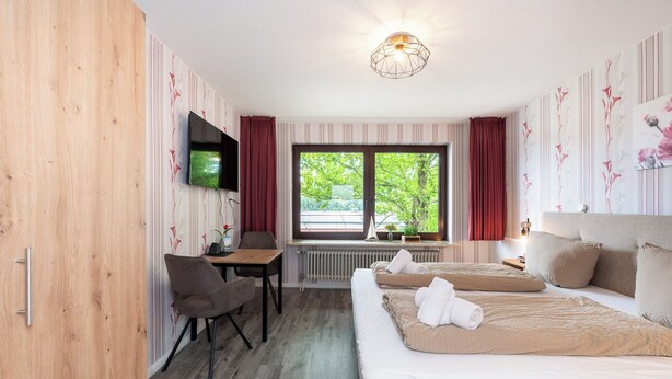 A hotel room with a wooden wardrobe, a desk with a chair, a TV mounted on the wall, a large window with curtains, and a bed with pillows. - Quartier 9 apartment in a quiet location and access to the sea time (Büsum)