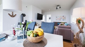 Un comedor con una mesa blanca, un cuenco de fruta y una jarra de agua. Se observa un televisor al fondo y un sofá en la sala.