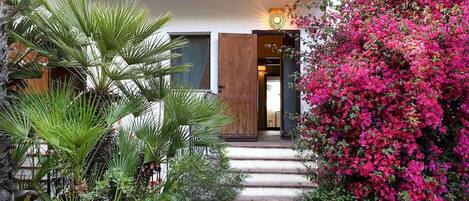 A house with a wooden door, a staircase leading to the entrance, and a garden with green plants and pink flowers.