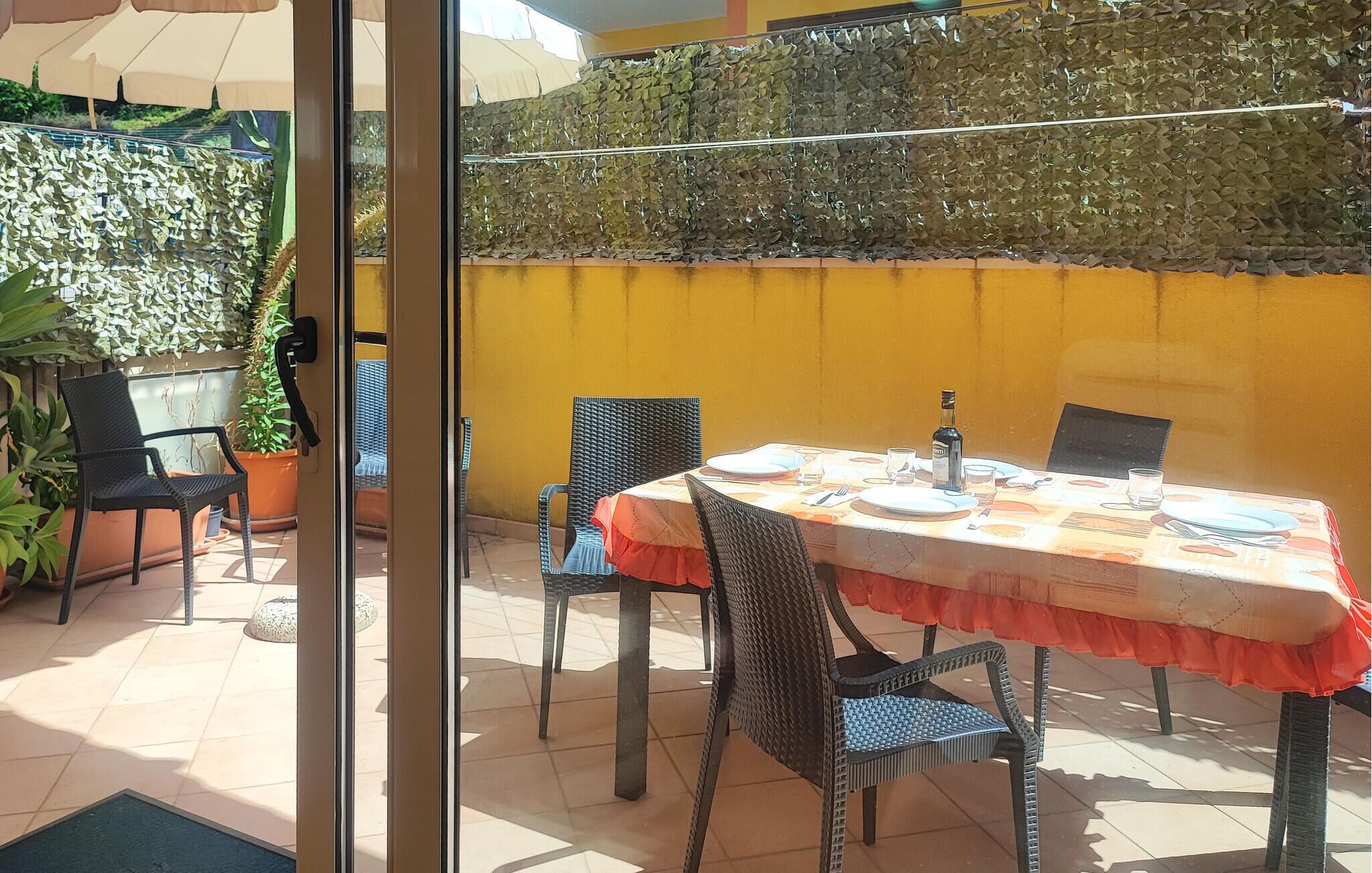 An outdoor patio with a table set for two, surrounded by wicker chairs, potted plants, and a yellow wall with a leafy green top.