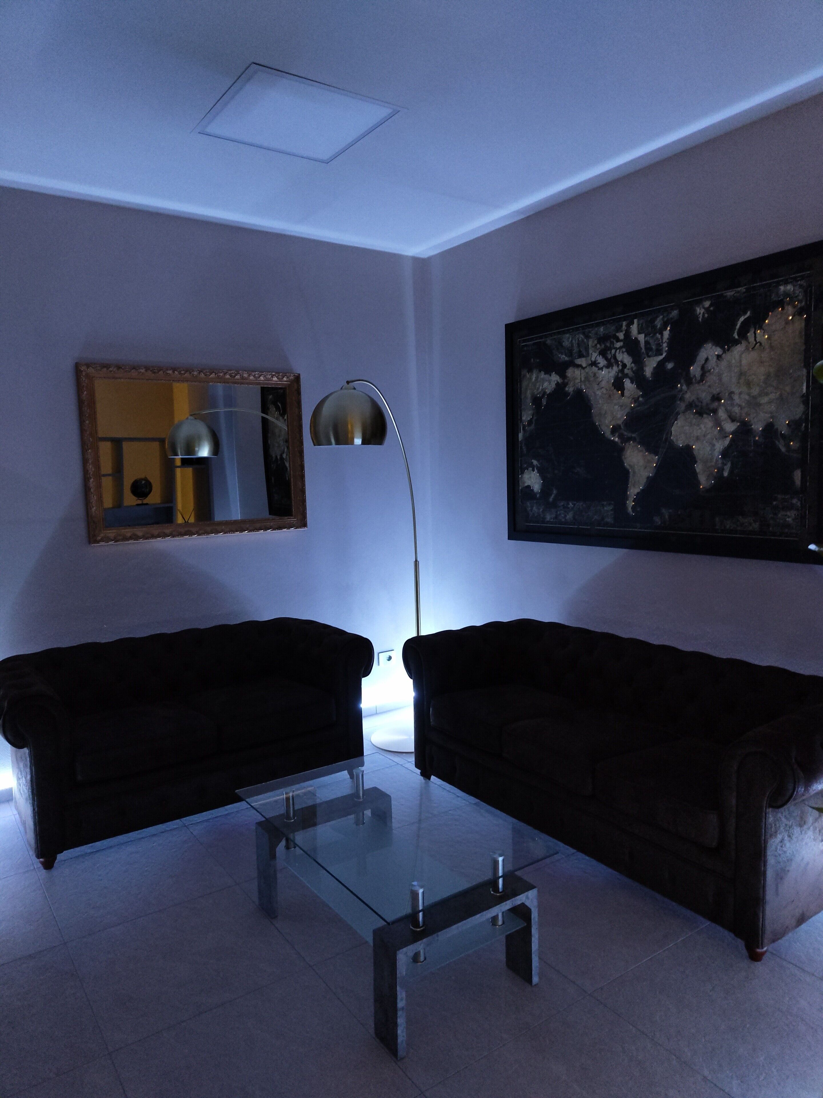Uma sala de estar moderna com dois sofás escuros, uma mesa de centro de vidro, um abajur, um espelho emoldurado e um grande quadro abstrato na parede.