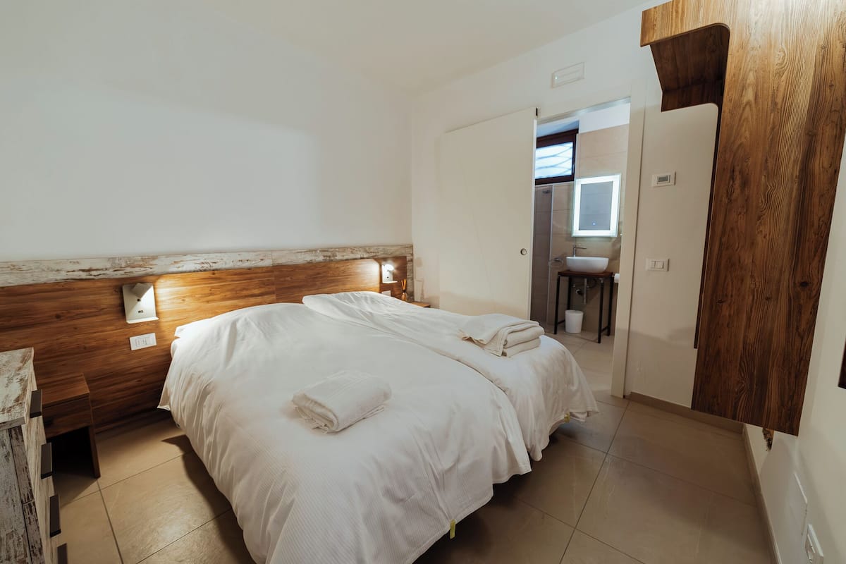 A double bed with white linens and a wooden headboard. There is a small bedside table with a lamp and a wall-mounted light. A wooden door and a bathroom with a sink and mirror are visible in the background.