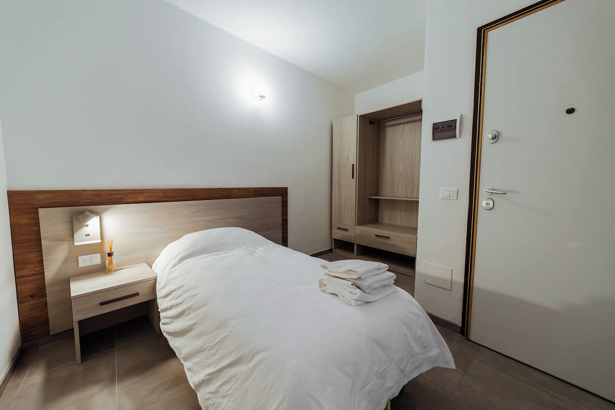 A neatly made bed with white linens, a bedside table with a lamp, and a wardrobe with a wooden finish.