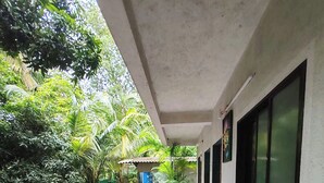A covered walkway with a tiled floor, flanked by lush greenery and a building with large windows. - Lotus Cottage (Alibag)
