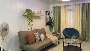 Ein Wohnzimmer mit einer Couch, einem runden Tisch, einem Sessel, einem Ventilator und einer Pflanze.