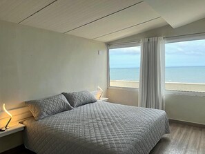 A bedroom with a bed, a nightstand, a lamp, and a window with a view of the sea.