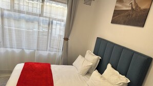 Una cama bien tendida con sábanas blancas y una manta roja, un cabecero tapizado en azul y un cuadro con jirafas en la pared.