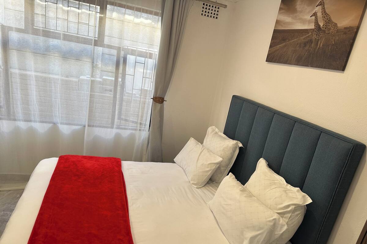 A neatly made bed with white linens and a red throw, a blue upholstered headboard, and a framed picture of giraffes on the wall.
