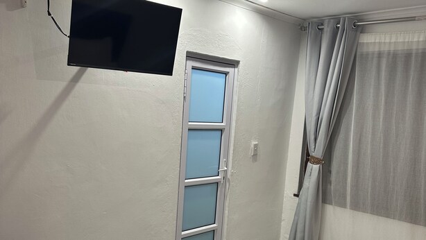 Un dormitorio con una cama, un televisor montado en la pared, una ventana con cortinas y una puerta con panel de vidrio esmerilado.