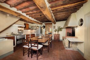 A rustic kitchen with exposed wooden beams, a stone fireplace, and a dining area with wooden chairs and a table.