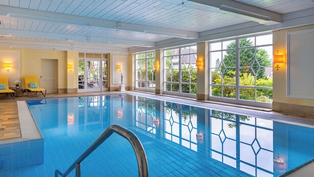 An indoor swimming pool with clear glass walls, wooden ceiling, and yellow chairs by the pool. - Relais & Châteaux Hotel Schwarzmatt (Badenweiler)