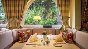 A dining area with a table set for two, a potted plant, and a window with curtains. - Relais & Châteaux Hotel Schwarzmatt (Badenweiler)