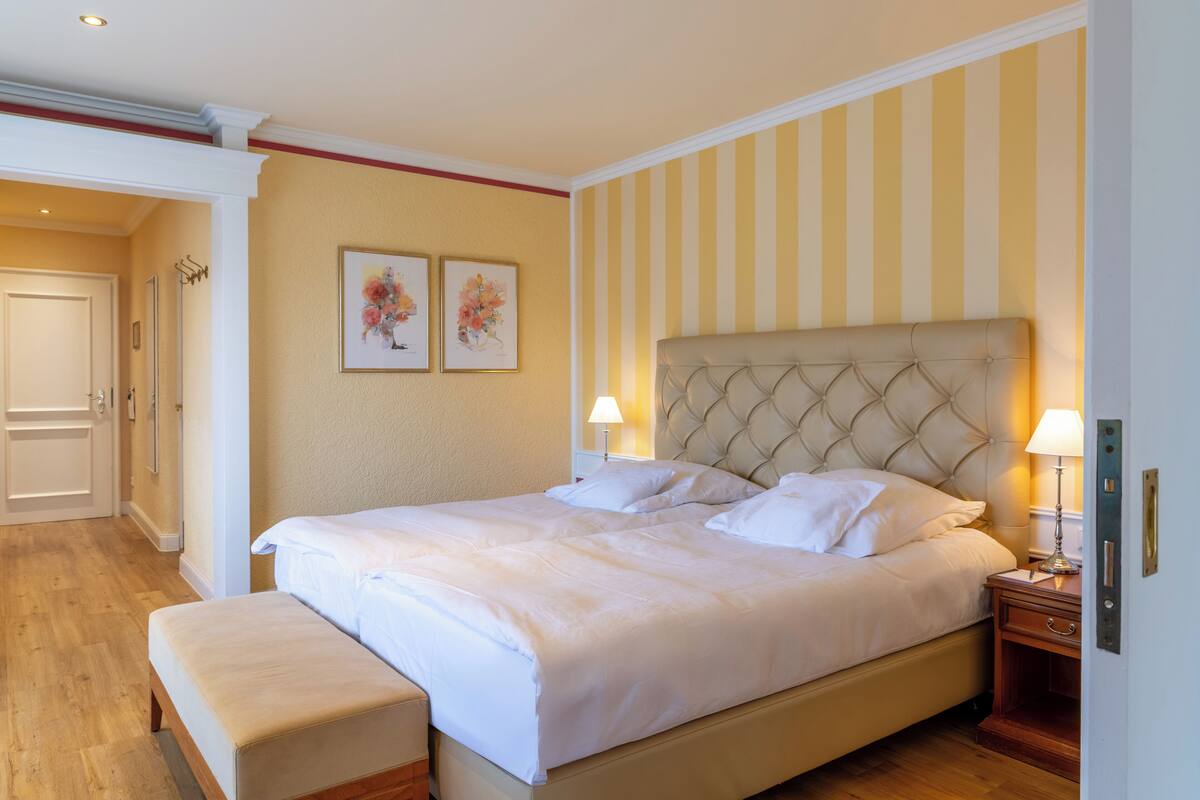 A hotel room with a large bed, bedside lamps, a bench, and two framed floral artworks on the wall.