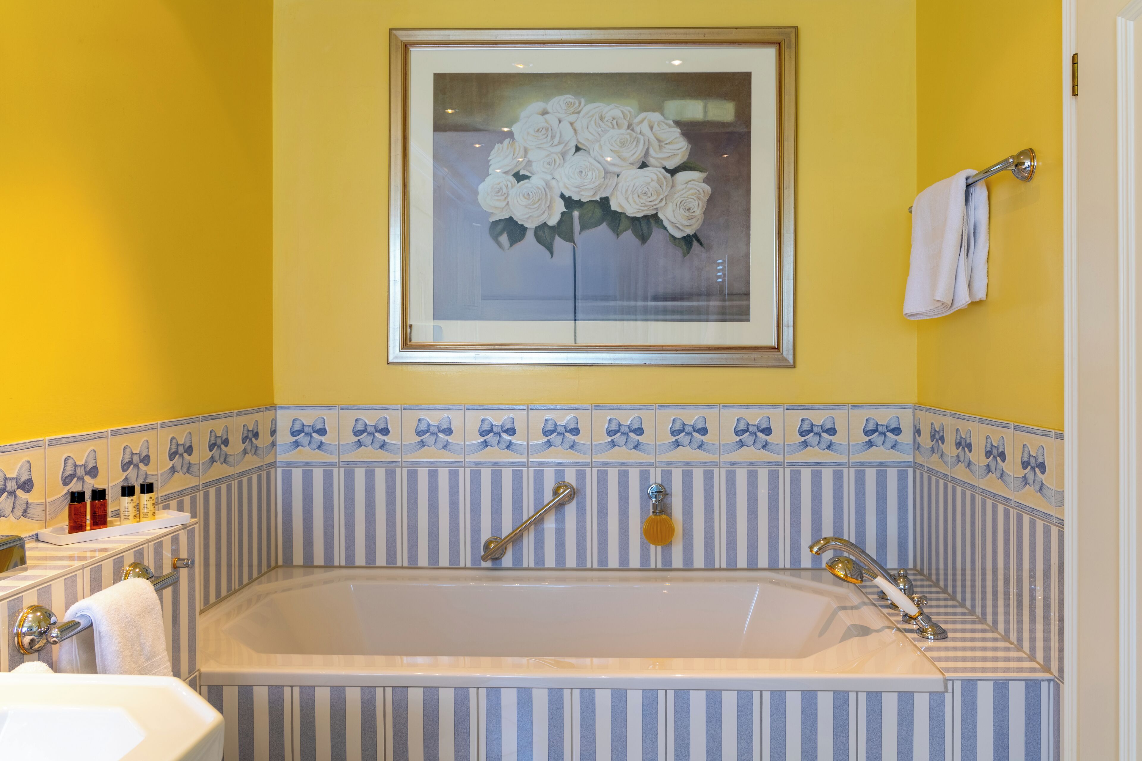 Ein Badezimmer mit einer Badewanne, Streifenfliesen, einem Fenster und einem floralen Bild in einem Rahmen sowie einem Handtuchhalter.