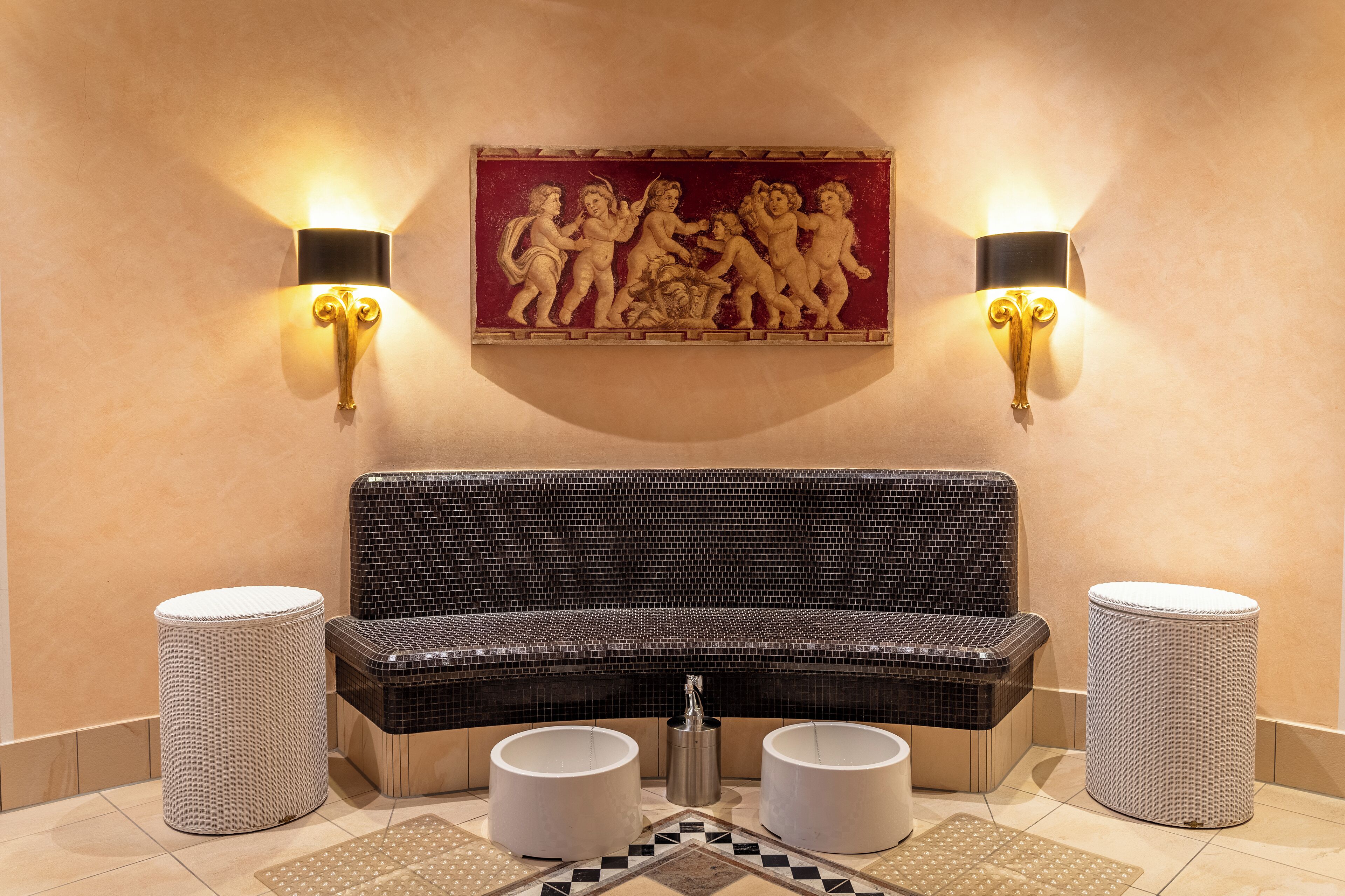 A lobby with a black cushioned bench, two wall-mounted sconces, a mosaic artwork, and two cylindrical stools.