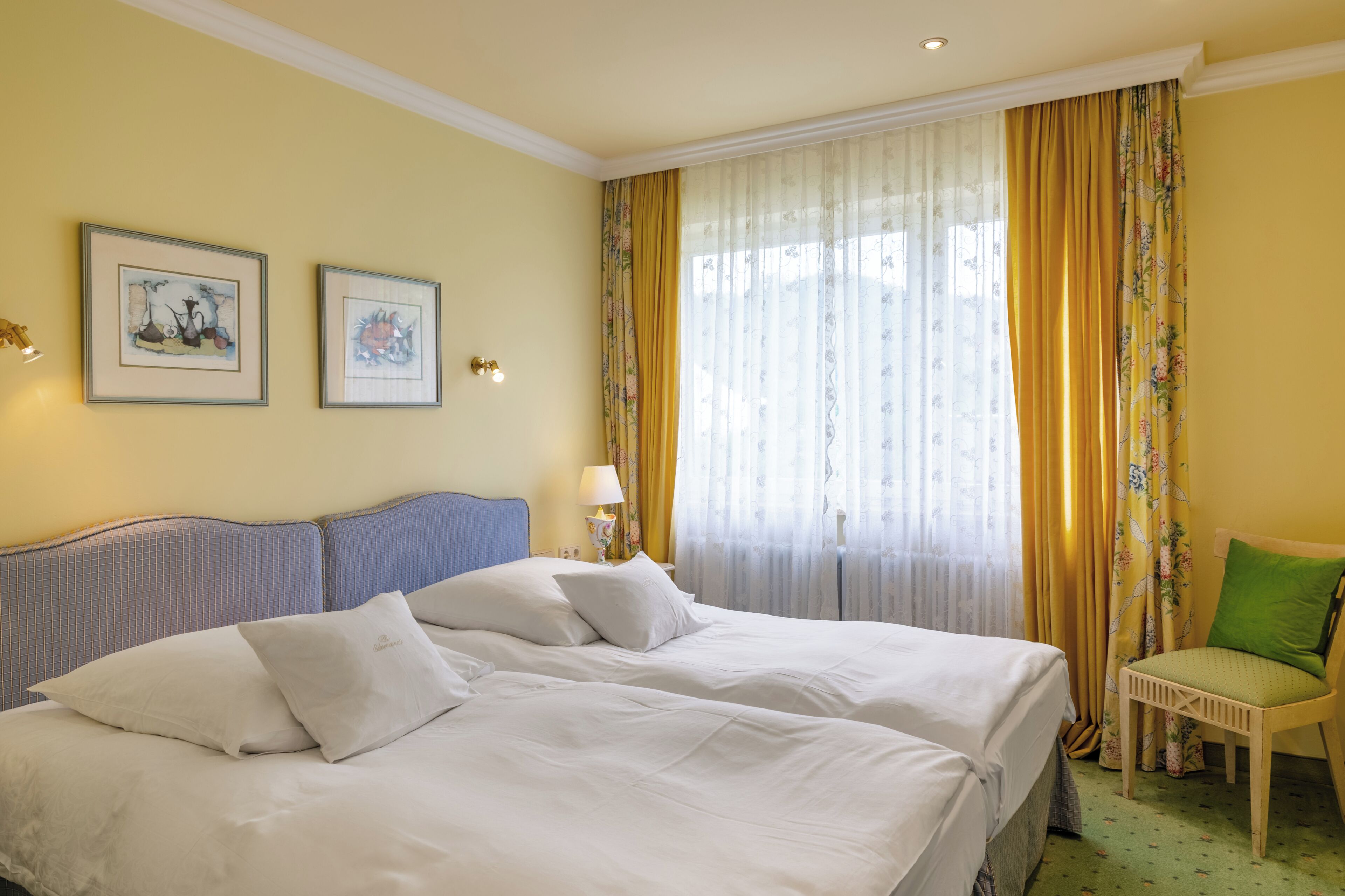 Ein Hotelzimmer mit zwei Betten, einem Sessel, einer Lampe und zwei gerahmten Bildern an der Wand.
