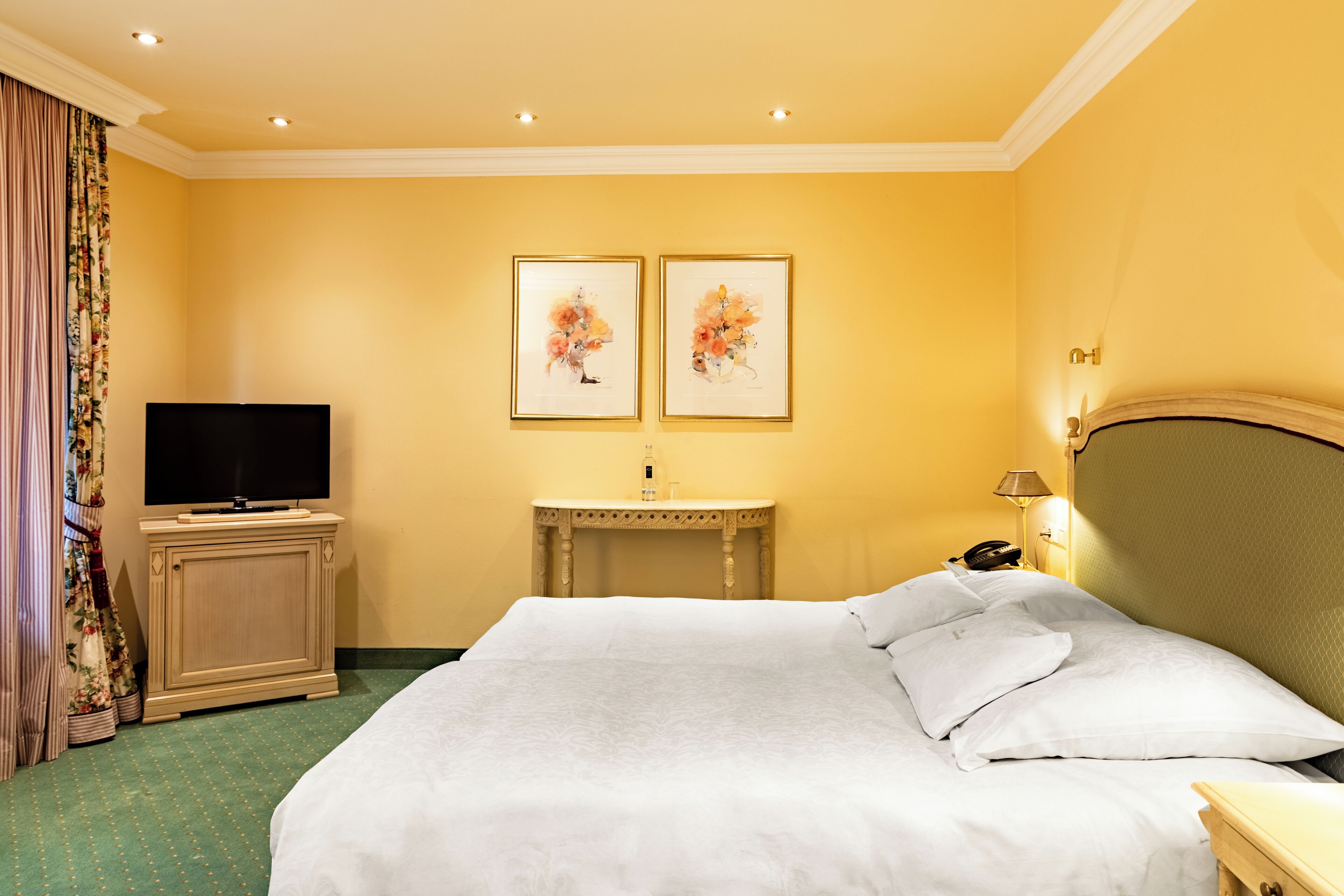 A hotel room with a bed, a television, a bedside table, a lamp, and two framed floral paintings on the wall.