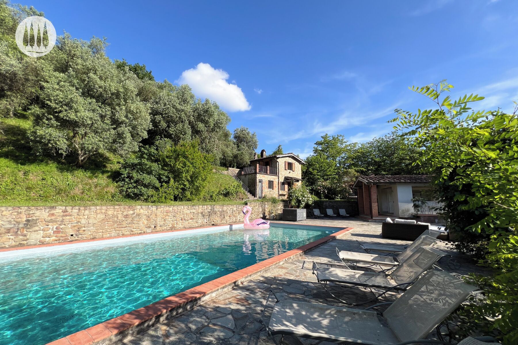 A swimming pool with lounge chairs, a stone wall, and a building with a balcony in the background.