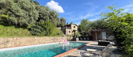 A swimming pool with lounge chairs, a stone wall, and a building with a balcony in the background.