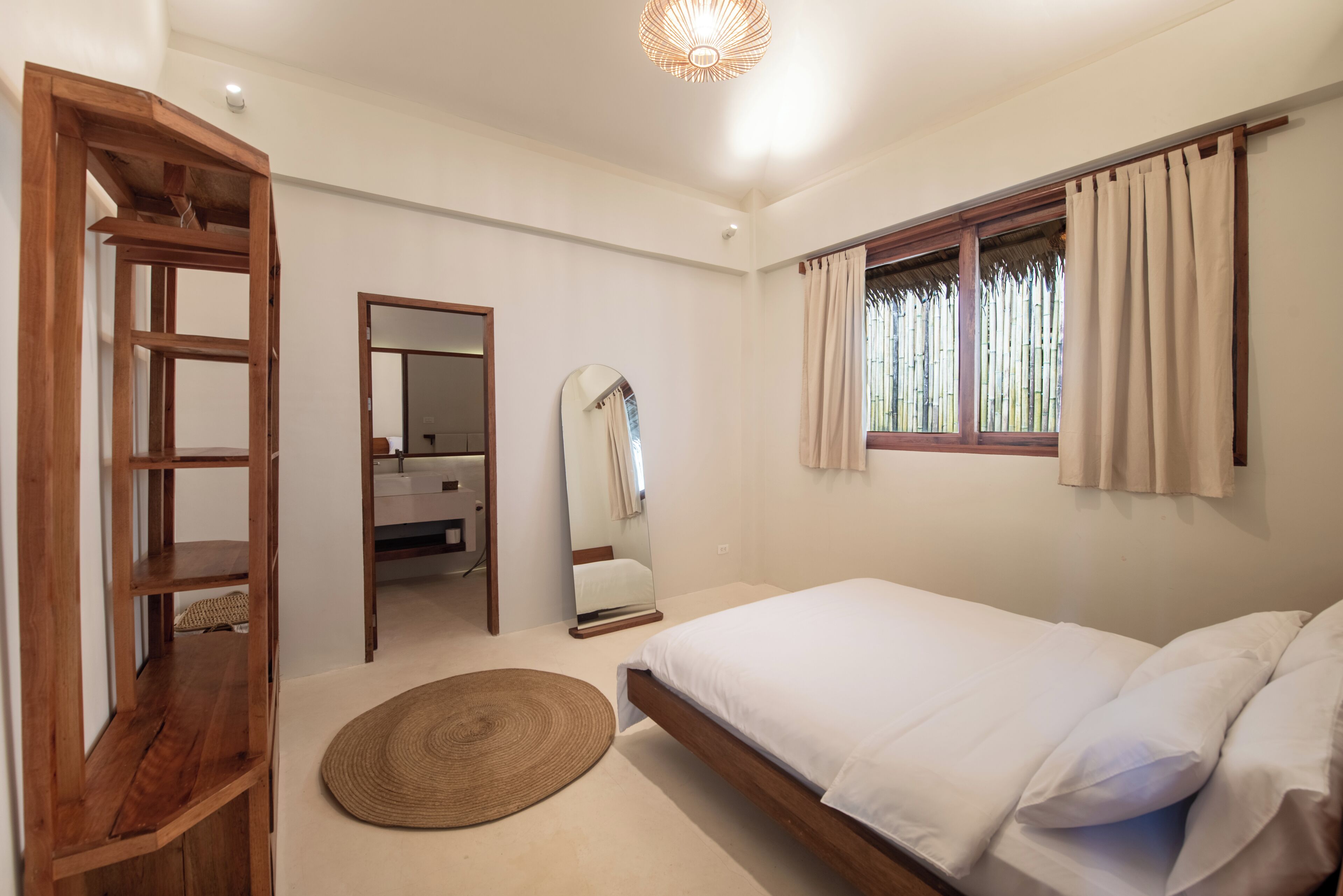 Une chambre avec un lit en bois, un tapis rond en fibres tressées, un miroir et une fenêtre avec des rideaux.