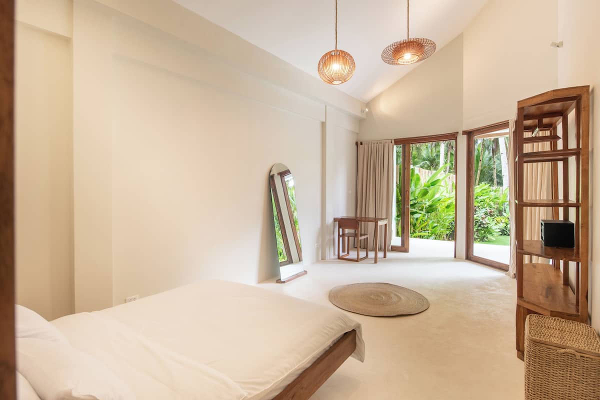 A bedroom with a bed, a round woven rug, a wooden chair, a table, and a large mirror.