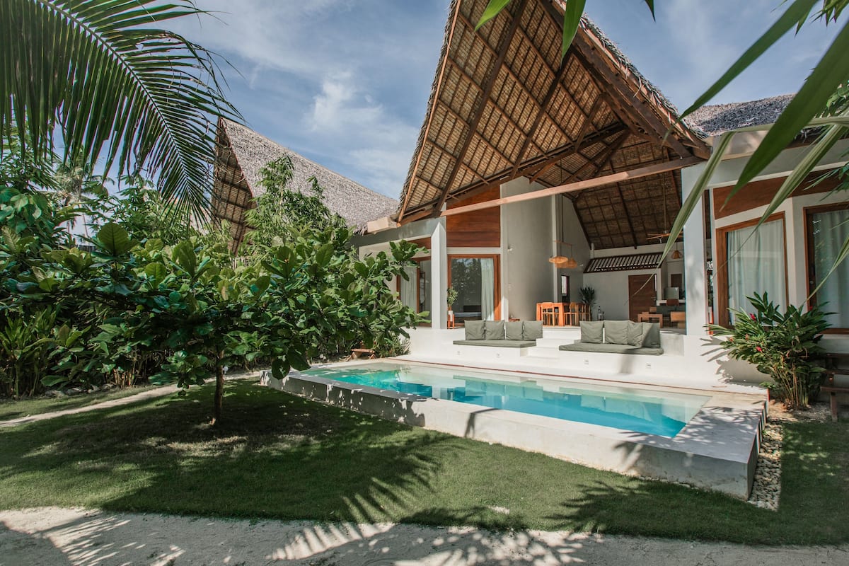 A modern house with a swimming pool, surrounded by lush greenery and a thatched roof structure.