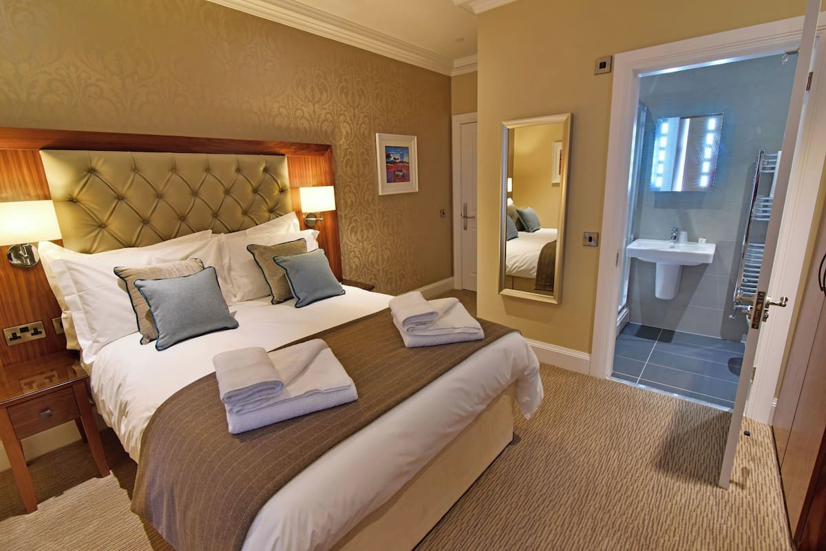 Une chambre d’hôtel avec un grand lit, une table de chevet, une commode, une salle de bain avec un lavabo et un miroir, et une porte donnant sur une autre pièce.