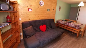 Ein gemütliches Wohnzimmer mit einer Holzwand, einem Sofa mit einem herzförmigen Kissen, einem Esstisch für vier Personen und einem kleinen Fernseher auf einem Regal.
