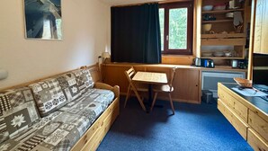Ein gemütliches Zimmer mit einer Holzdavonia, einem kleinen Tisch, einem Fenster mit Vorhängen, einer Küchenzeile mit Holdelementen und einem Bild an der Wand.