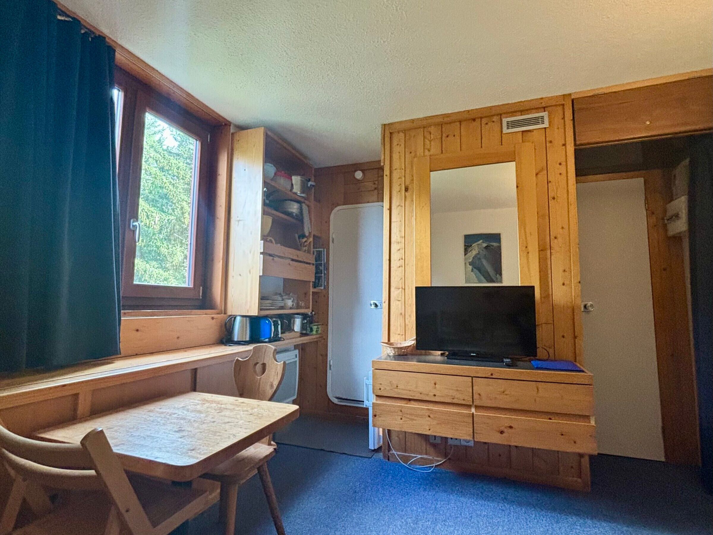 Une pièce de taille modeste, comprenant un bureau en bois, une chaise, un téléviseur et un réfrigérateur.