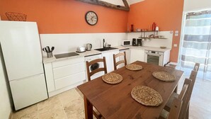 Een kleine keuken met witte kastjes, een houten tafel met gevlochten placemats en een klok aan de muur.