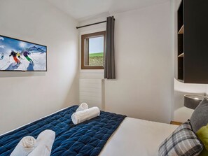 A bedroom with a bed, a window with curtains, a radiator, a TV on the wall, and a shelf with a lamp. - Charming T2 apartment in Les Saisies, near slopes (Les Saisies)