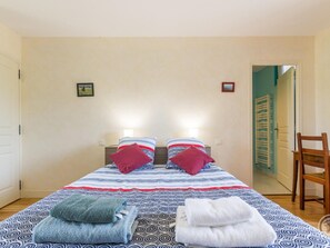Un dormitorio con una cama, dos almohadas, dos lámparas de noche, una silla de madera y una puerta que da a otra habitación.
